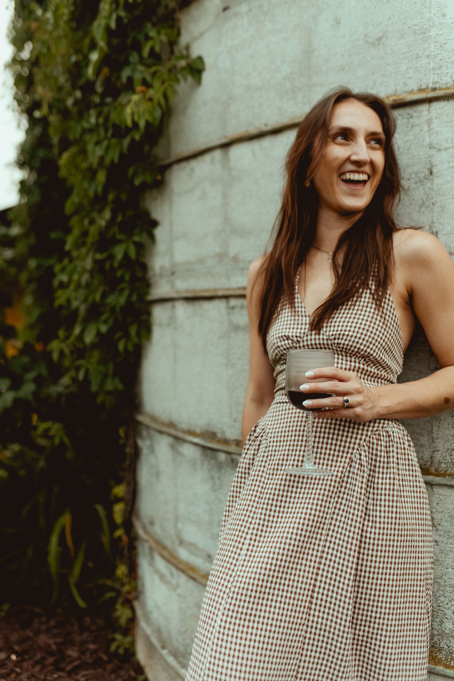 A laughing woman in a checkered sundress holds a drink while leaning against a concrete wall outdoors.