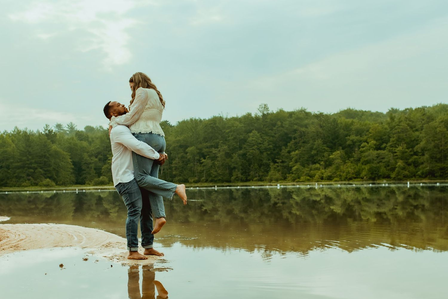 A man lifts a woman joyfully by a reflective lake surrounded by lush green trees on a cloudy day.