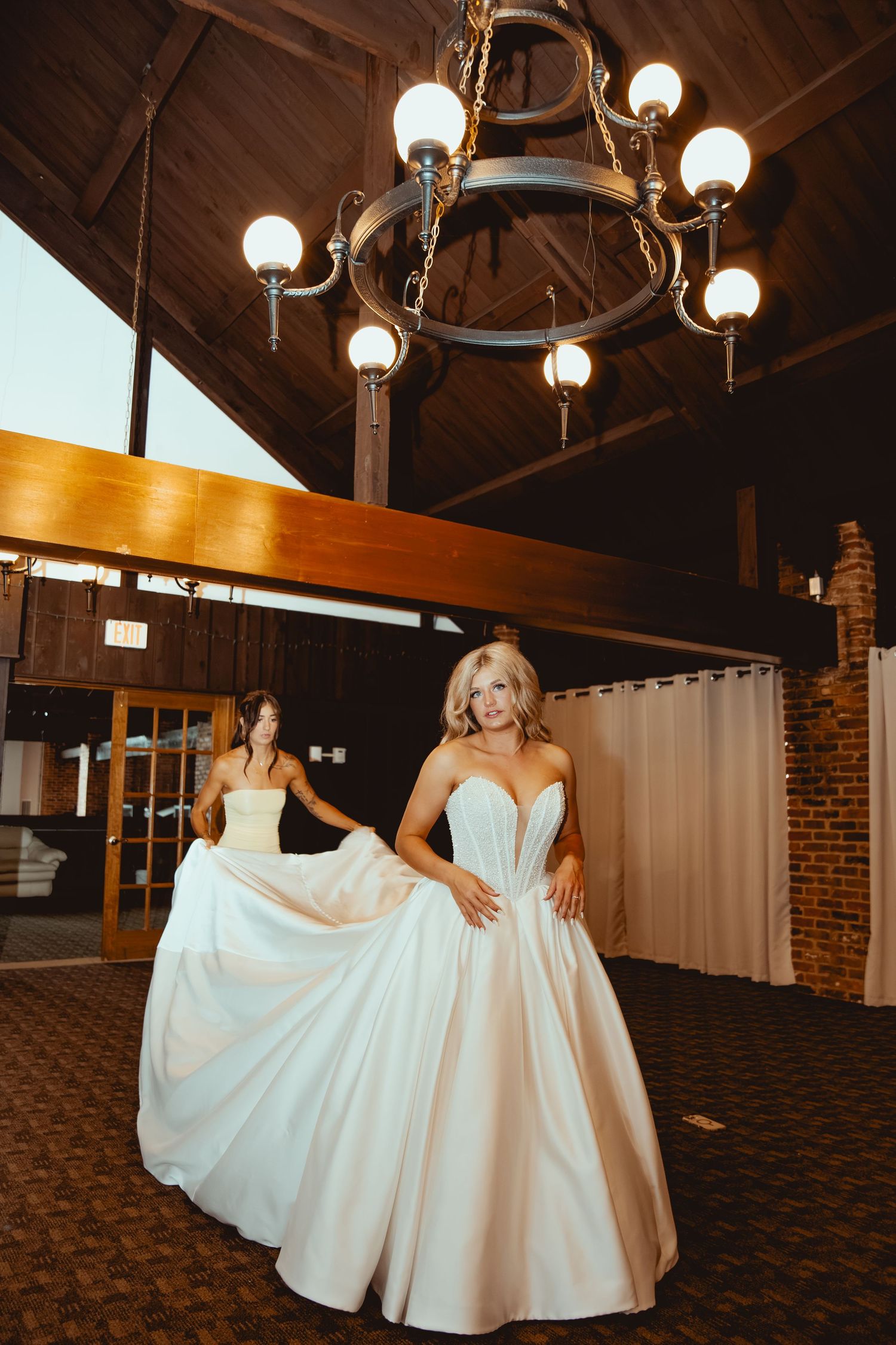 Bride in a strapless ball gown twirls in a rustic venue with chandelier while bridesmaid holds her train.