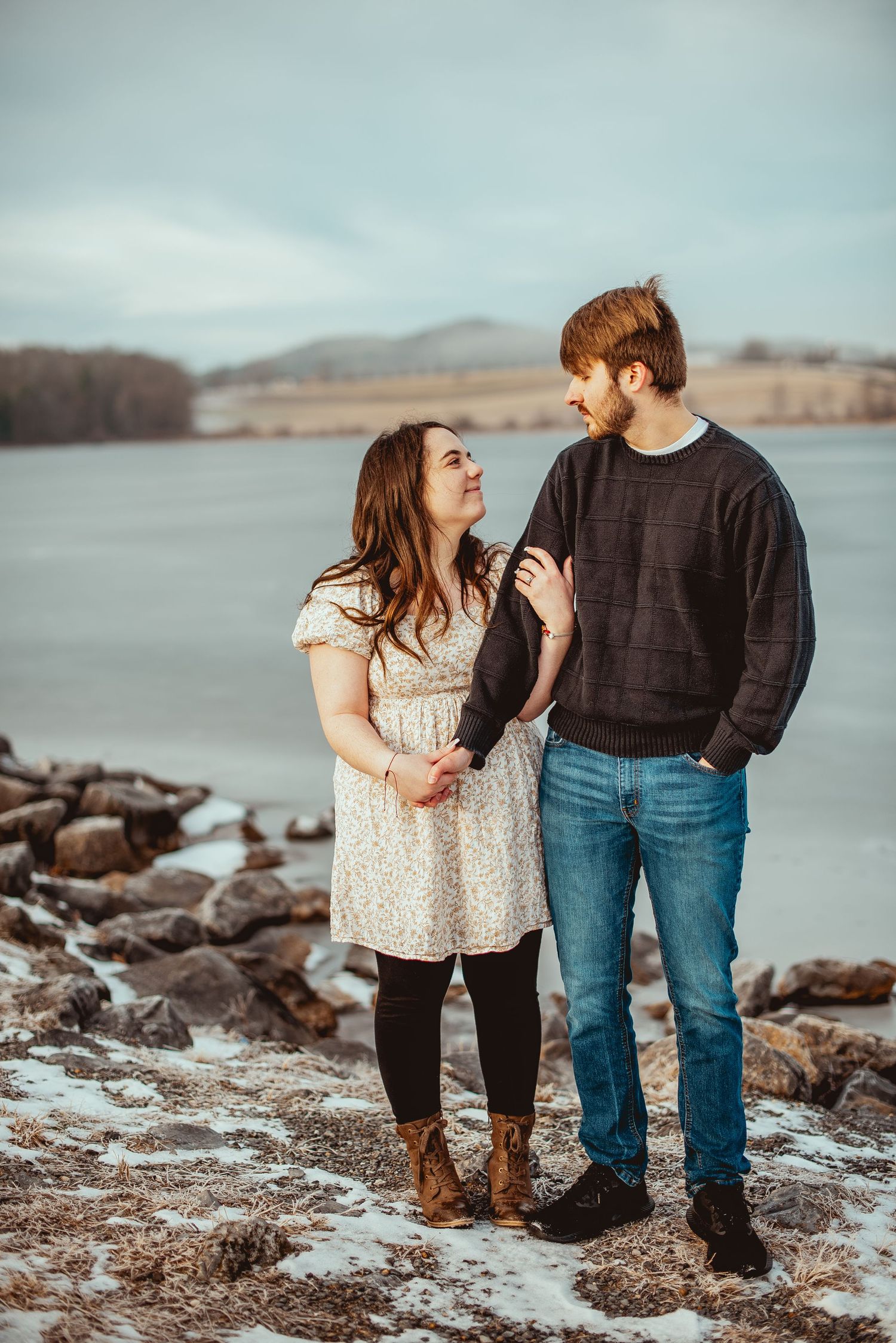 A couple shares a laugh while standing on a rocky shoreline with mountains in the background.