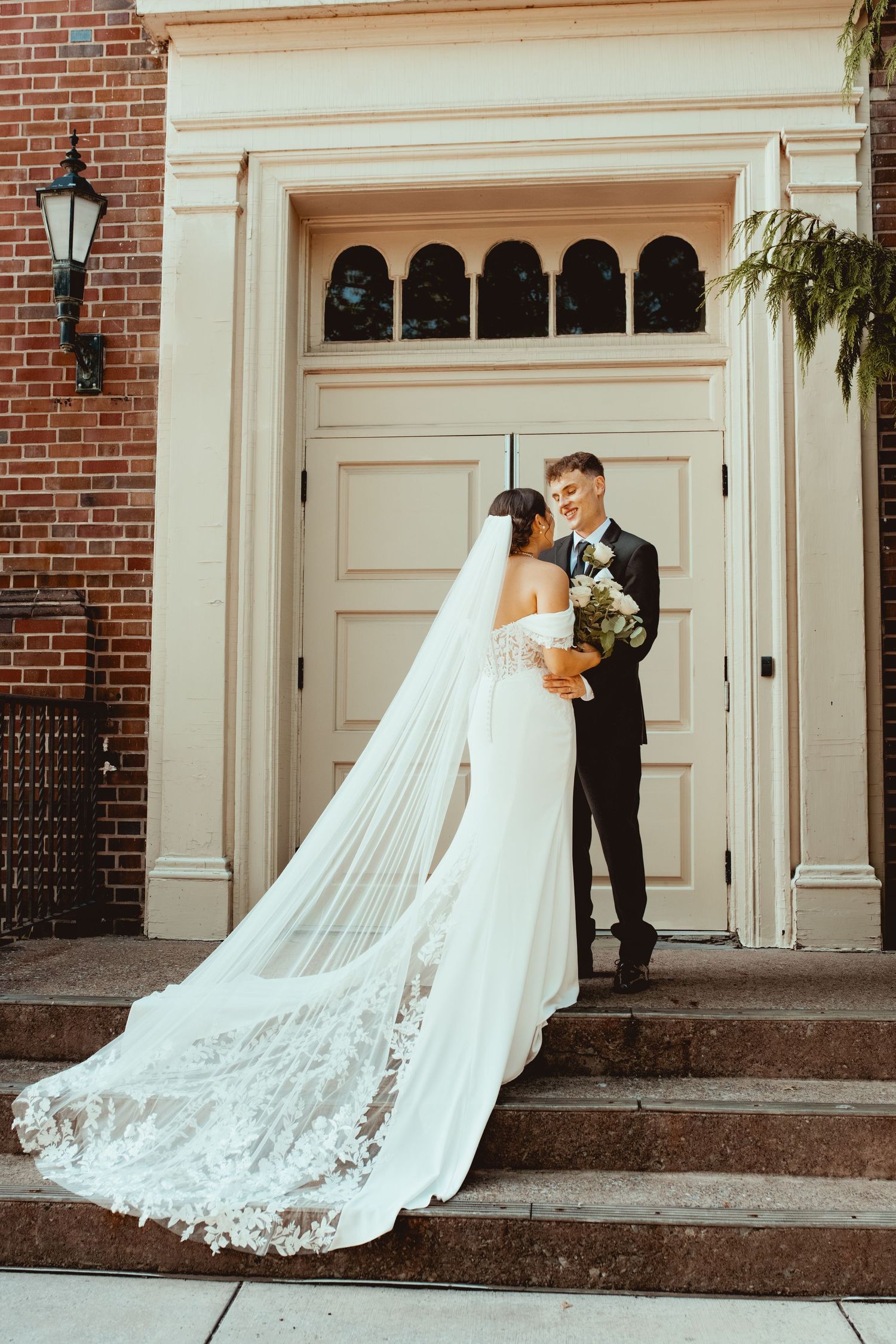 Romantic wedding couple poses in front of brick church entrance with flowing lace cathedral veil trailing behind.