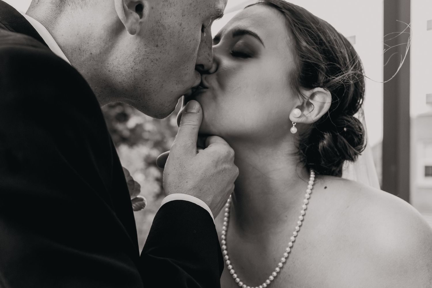 A romantic black and white wedding photo showing a tender kiss between a couple wearing formal attire and pearl jewelry.