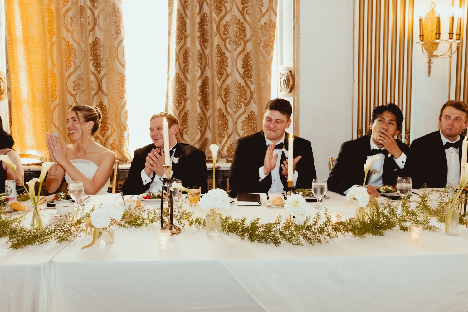 Wedding party seated at an elegant head table with gold decor and white floral arrangements.