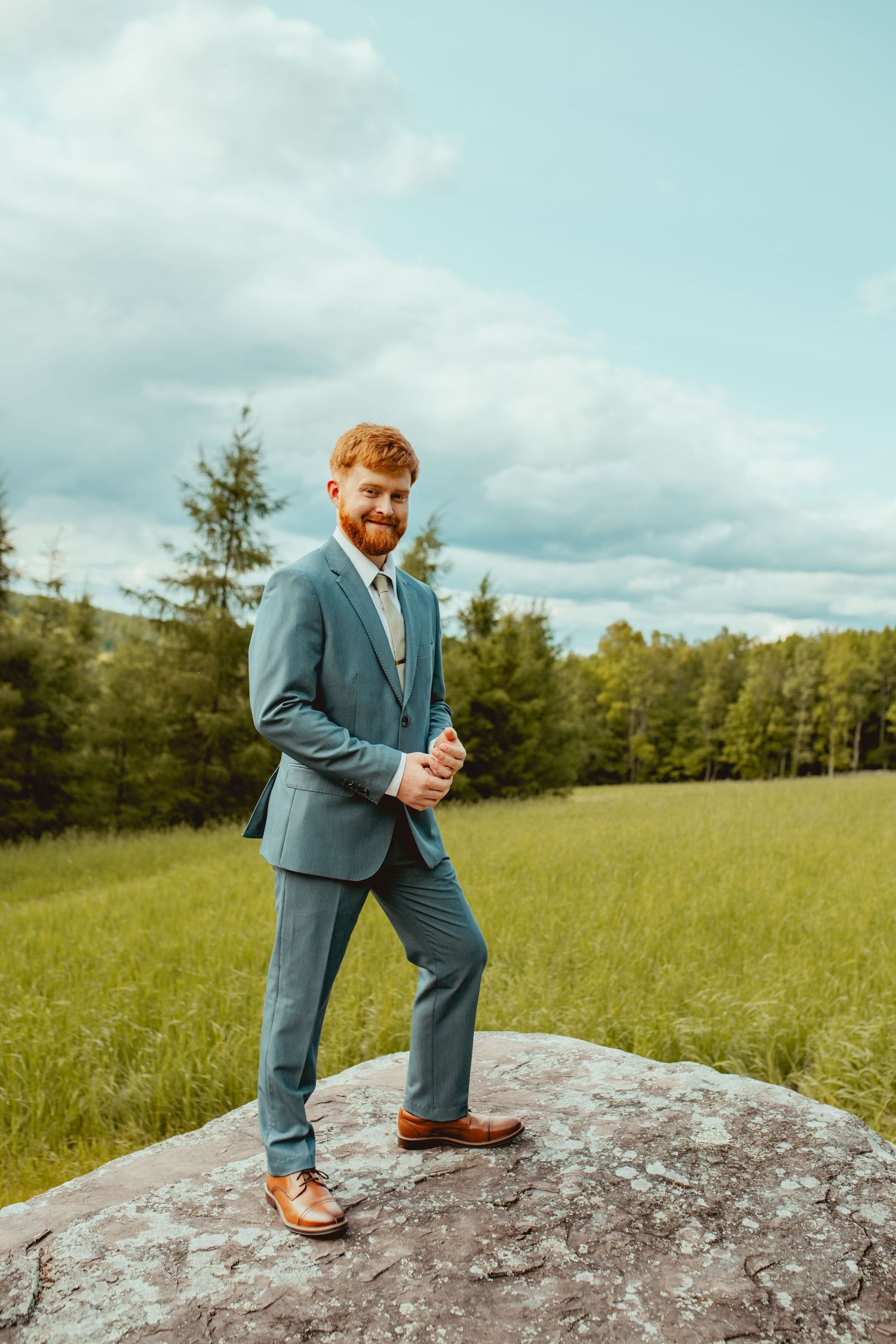 A person in a blue suit poses confidently on a large rock formation with evergreen trees in the background.
