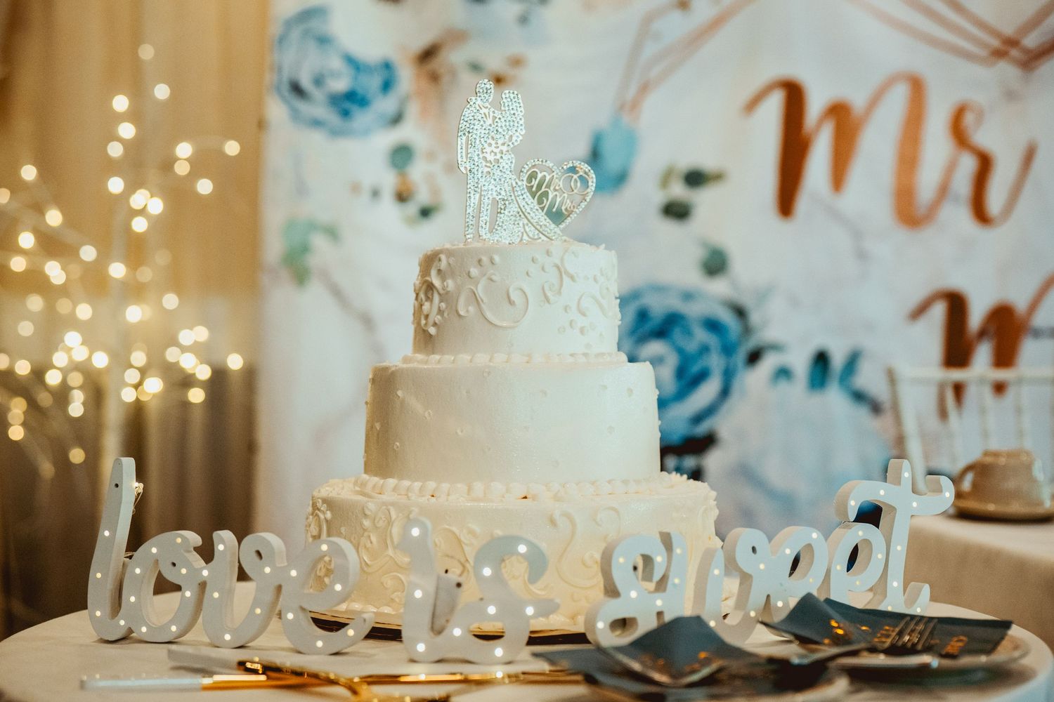 White wedding cake with delicate lace details and pearls displayed at an elegant reception with twinkling lights.