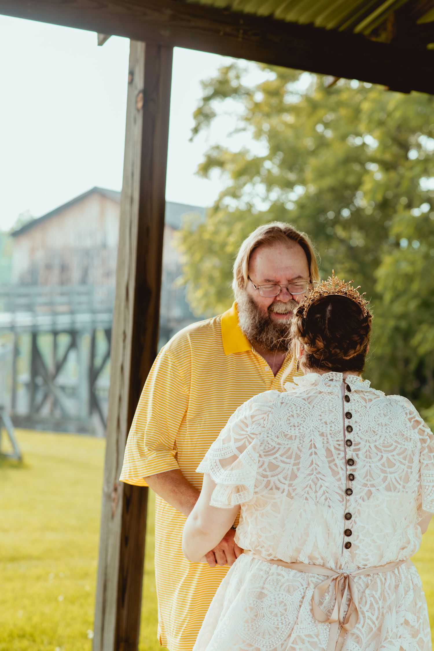 A couple embraces during their wedding celebration under a covered outdoor venue with sunny fields in background.