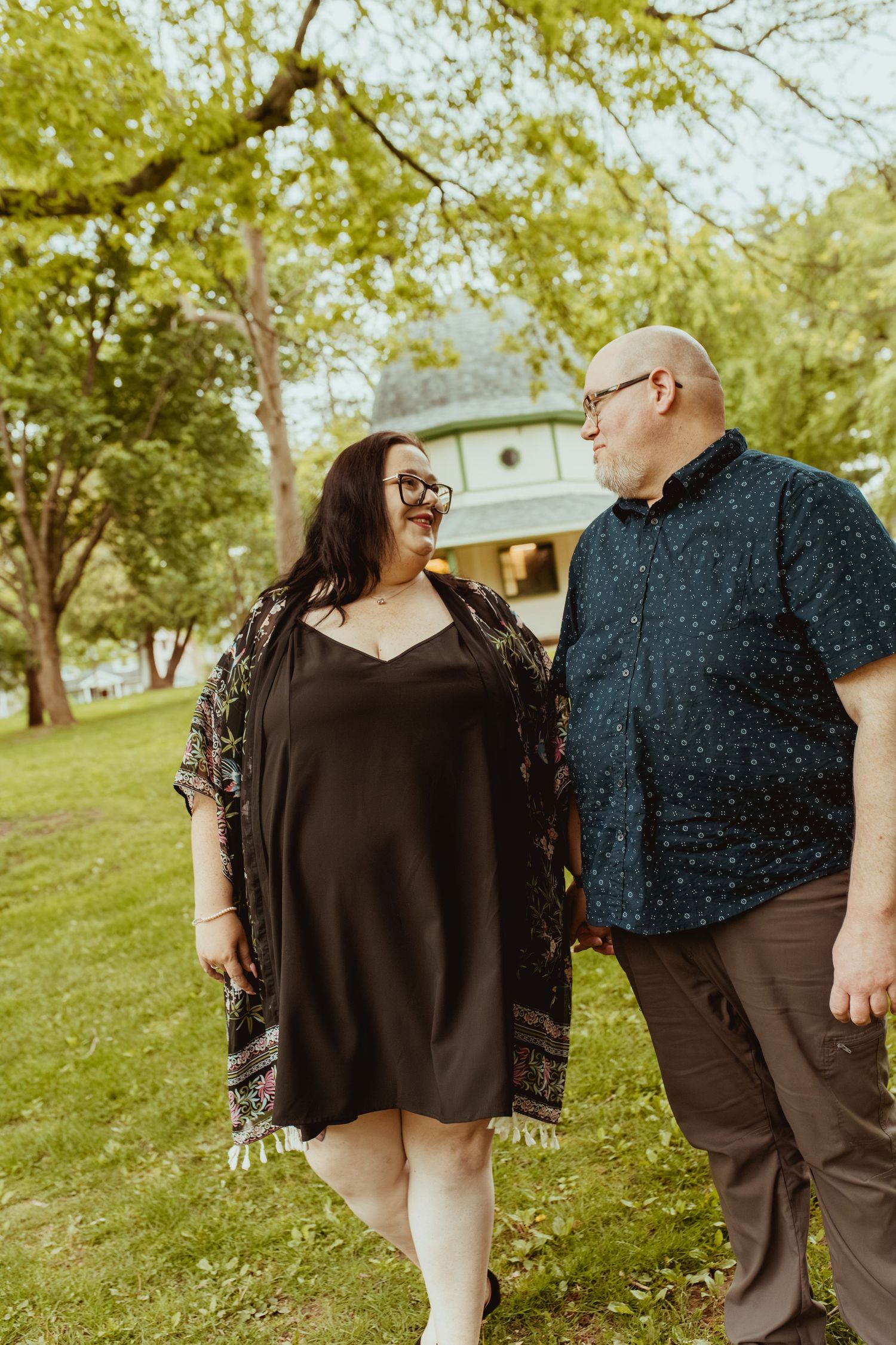 Couple walking hand in hand through a lush green park, smiling at each other.