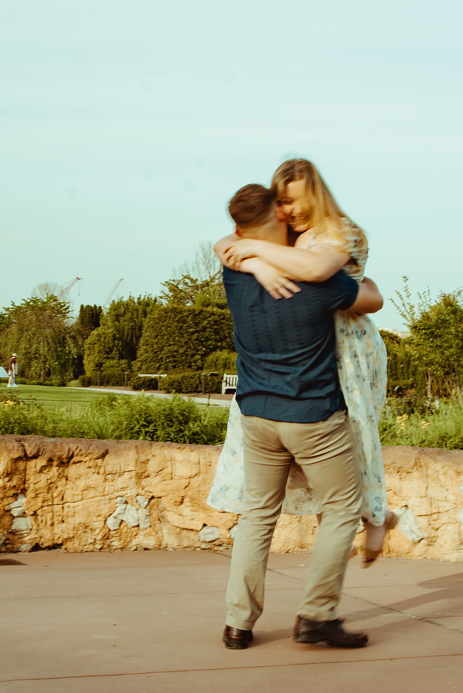 A couple embracing and spinning joyfully outdoors near a stone wall with lush green trees in the background.