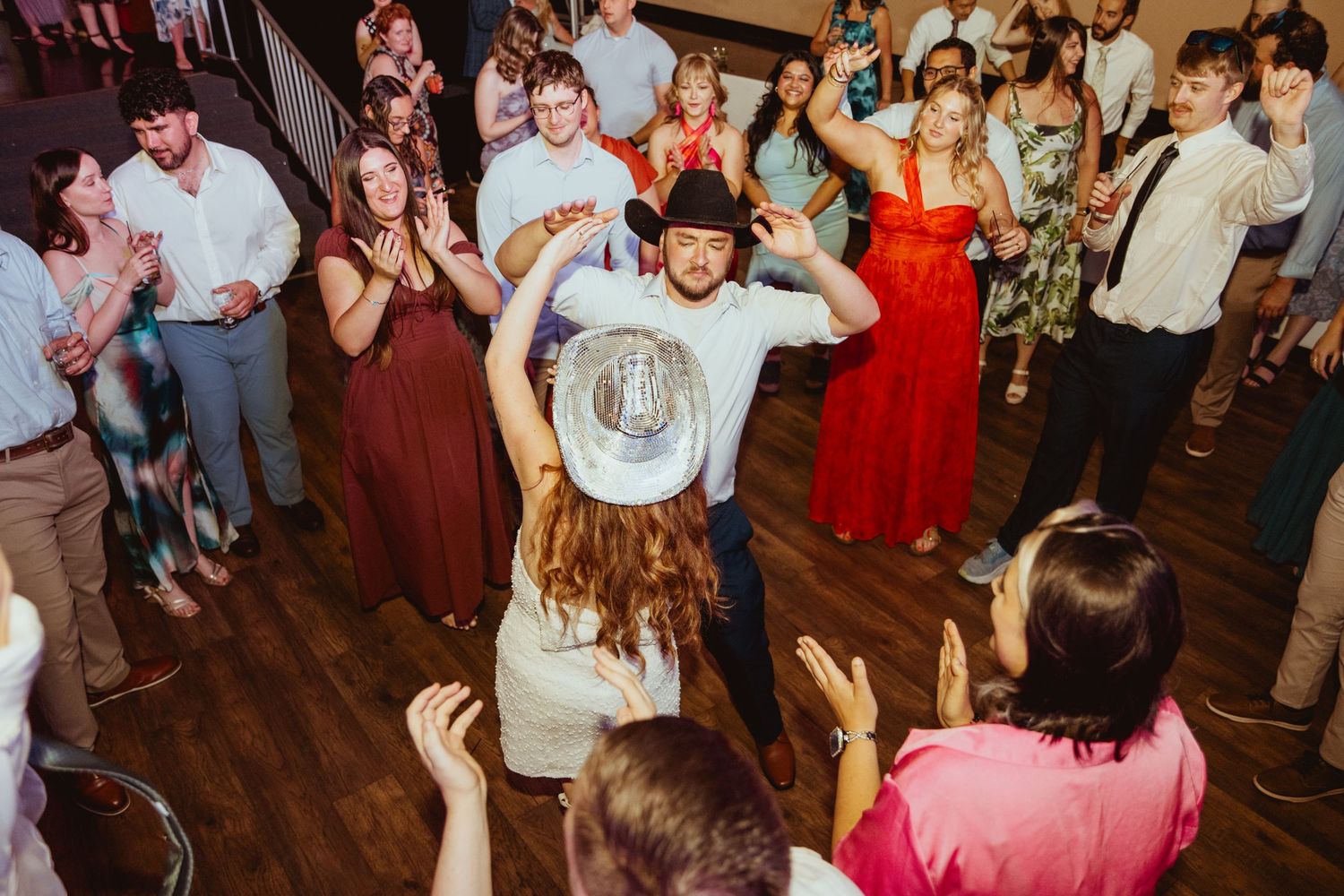 Guests dancing at a lively wedding reception, with a man in a cowboy hat at the center of the dance floor.