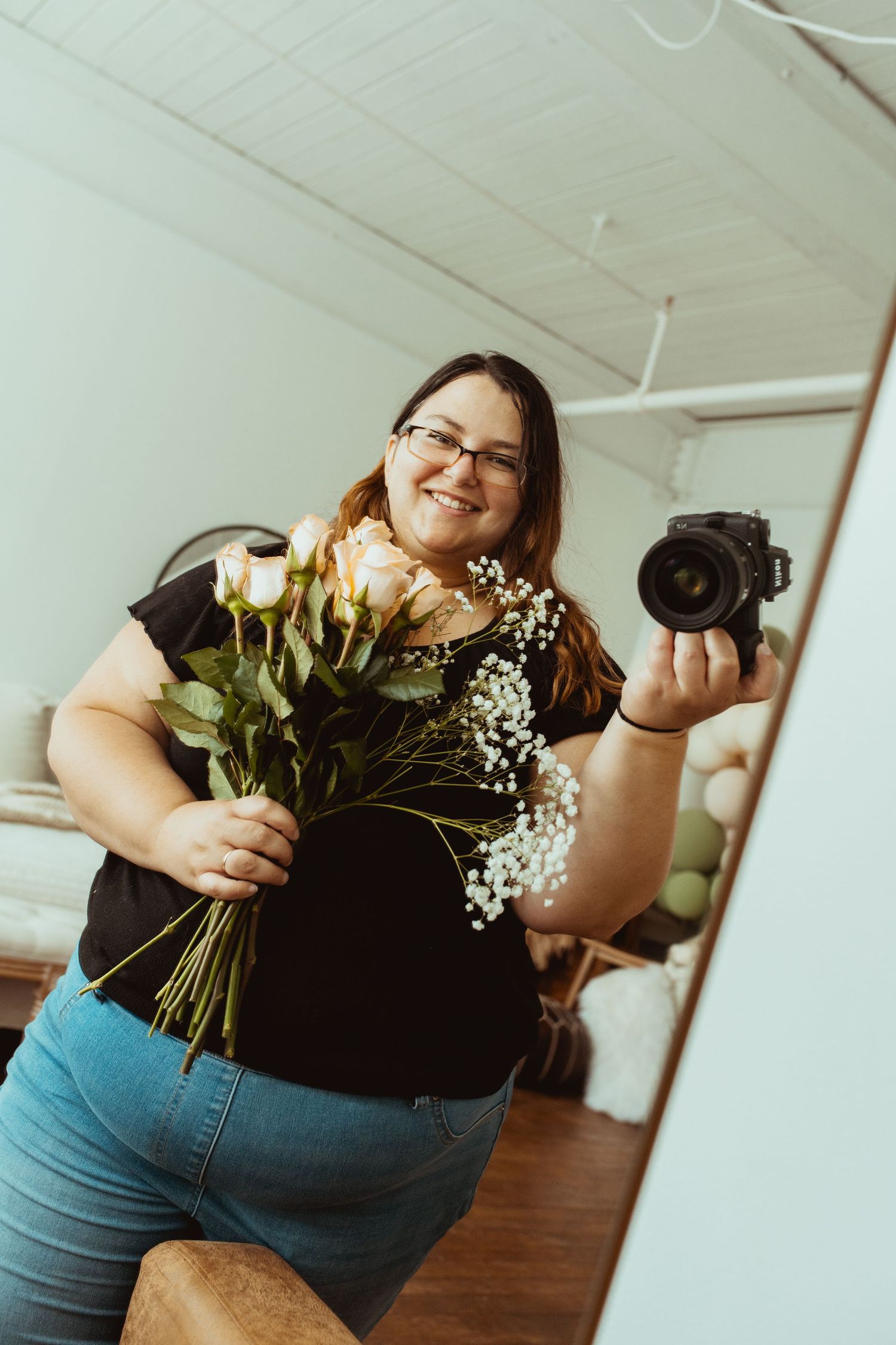 Woman smiling holding roses and baby's breath bouquet while taking mirror selfie with DSLR camera indoors.