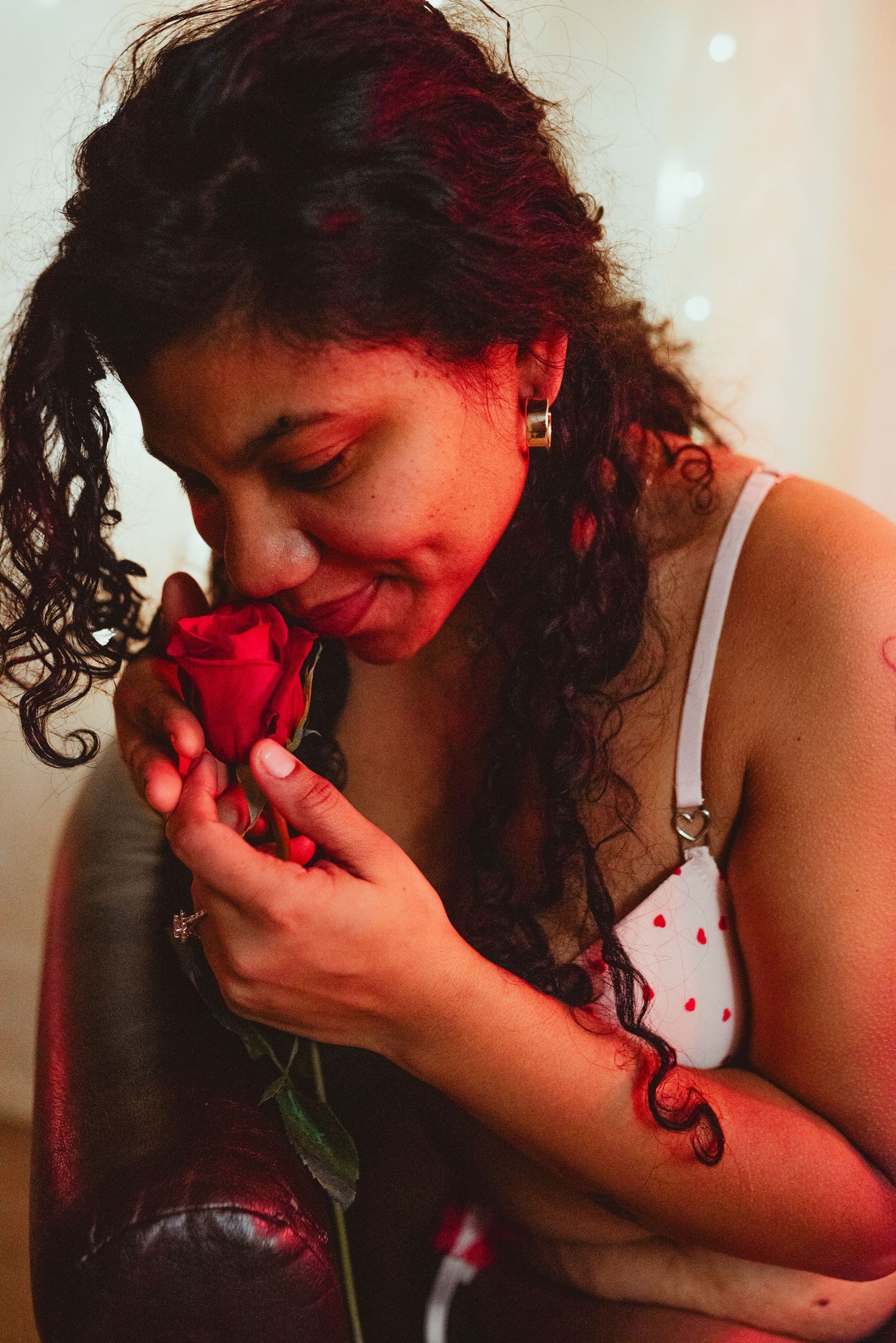 Woman with curly hair smelling a red rose, wearing a white floral top, in a warm romantic setting.