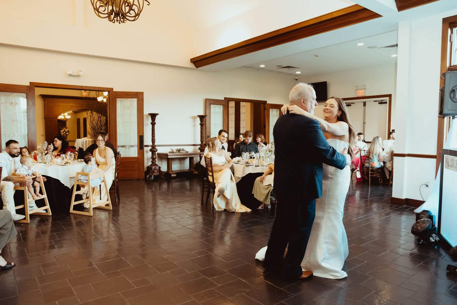 Bride and groom share their first dance at elegant wedding reception with guests seated at decorated tables in background.