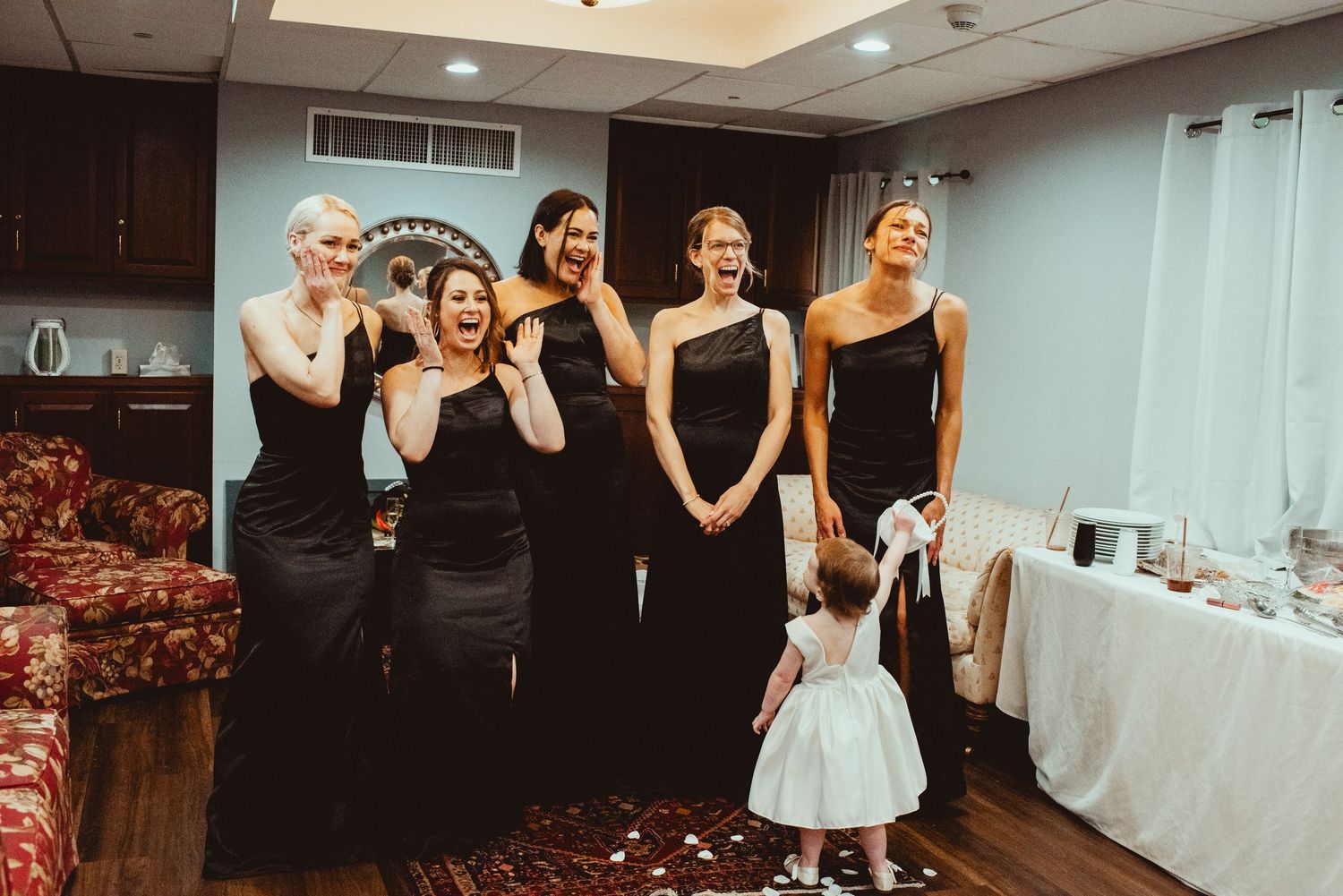 Bridesmaids in black dresses sharing a joyful moment with flower girl in white dress.