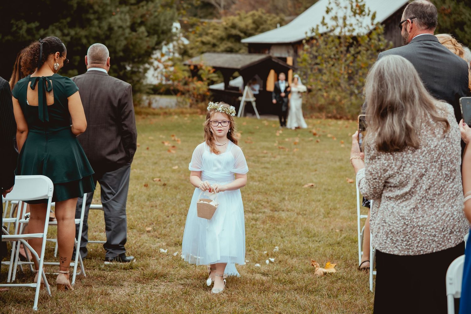 Wedding guests gather on a sunny autumn day during an outdoor ceremony with colorful fall foliage in the background.