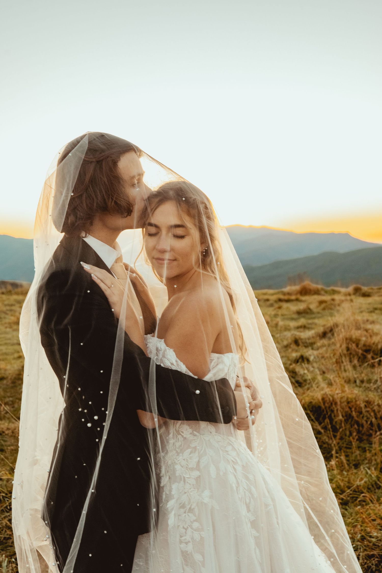 Intimate moment between newlyweds under bridal veil with mountains and sunset in background.