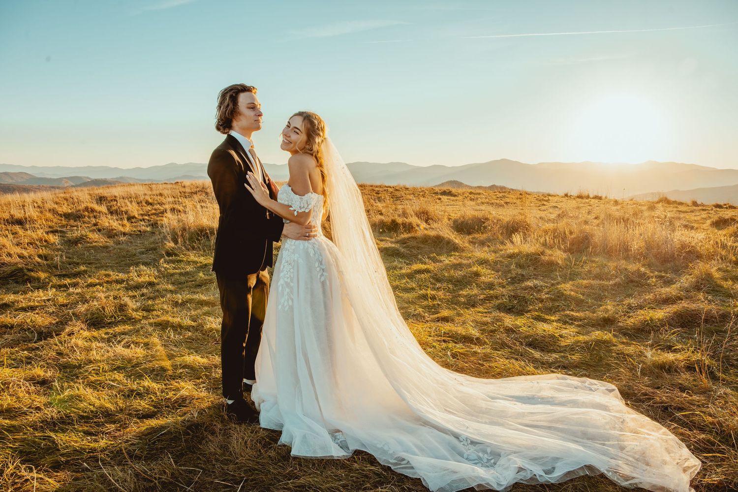 Newlywed couple embracing on hilltop during golden sunset with bride's long veil flowing in wind.