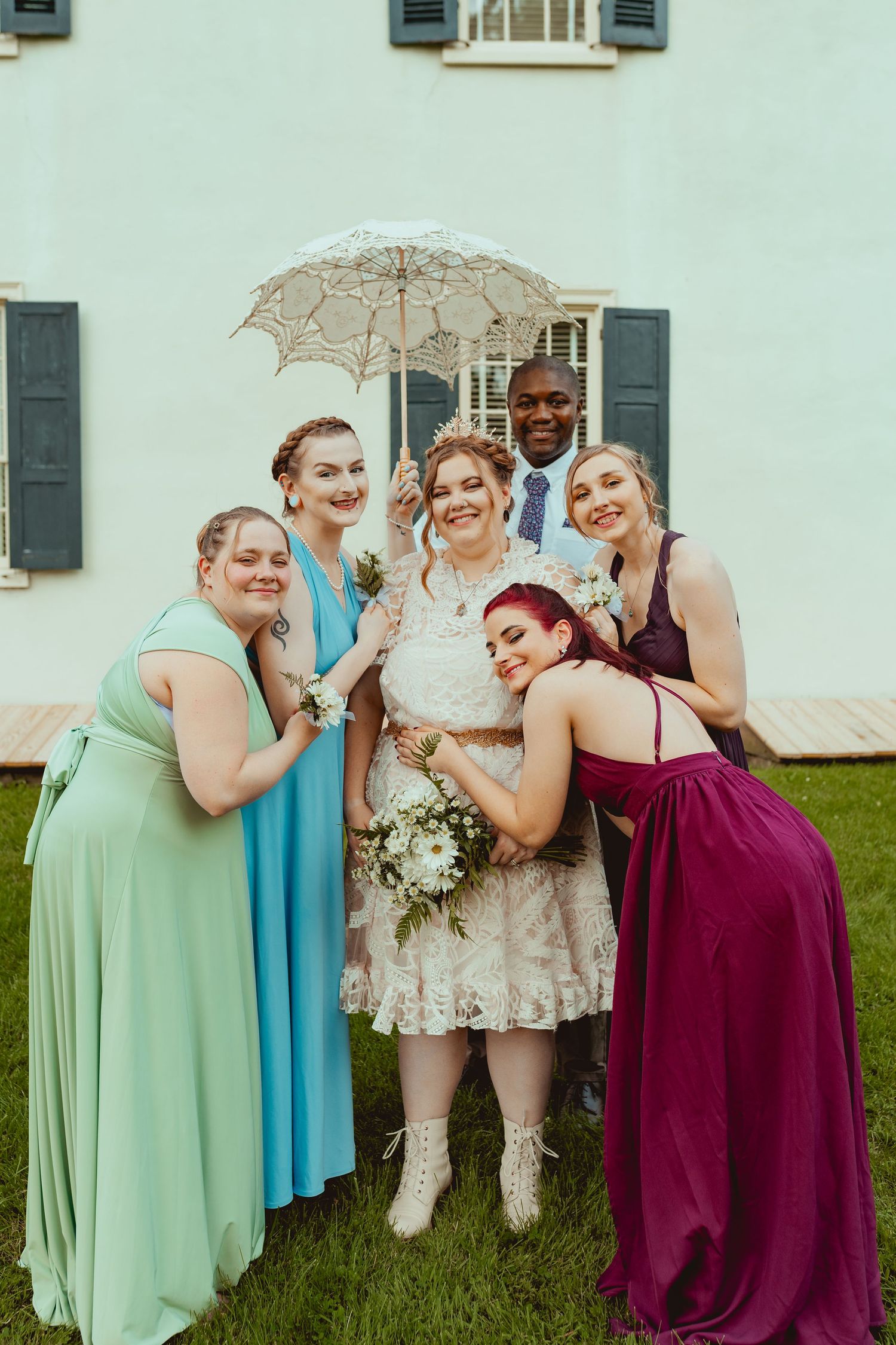 Bridesmaids in mint green, turquoise, and burgundy dresses pose playfully together on grass with a lace parasol.