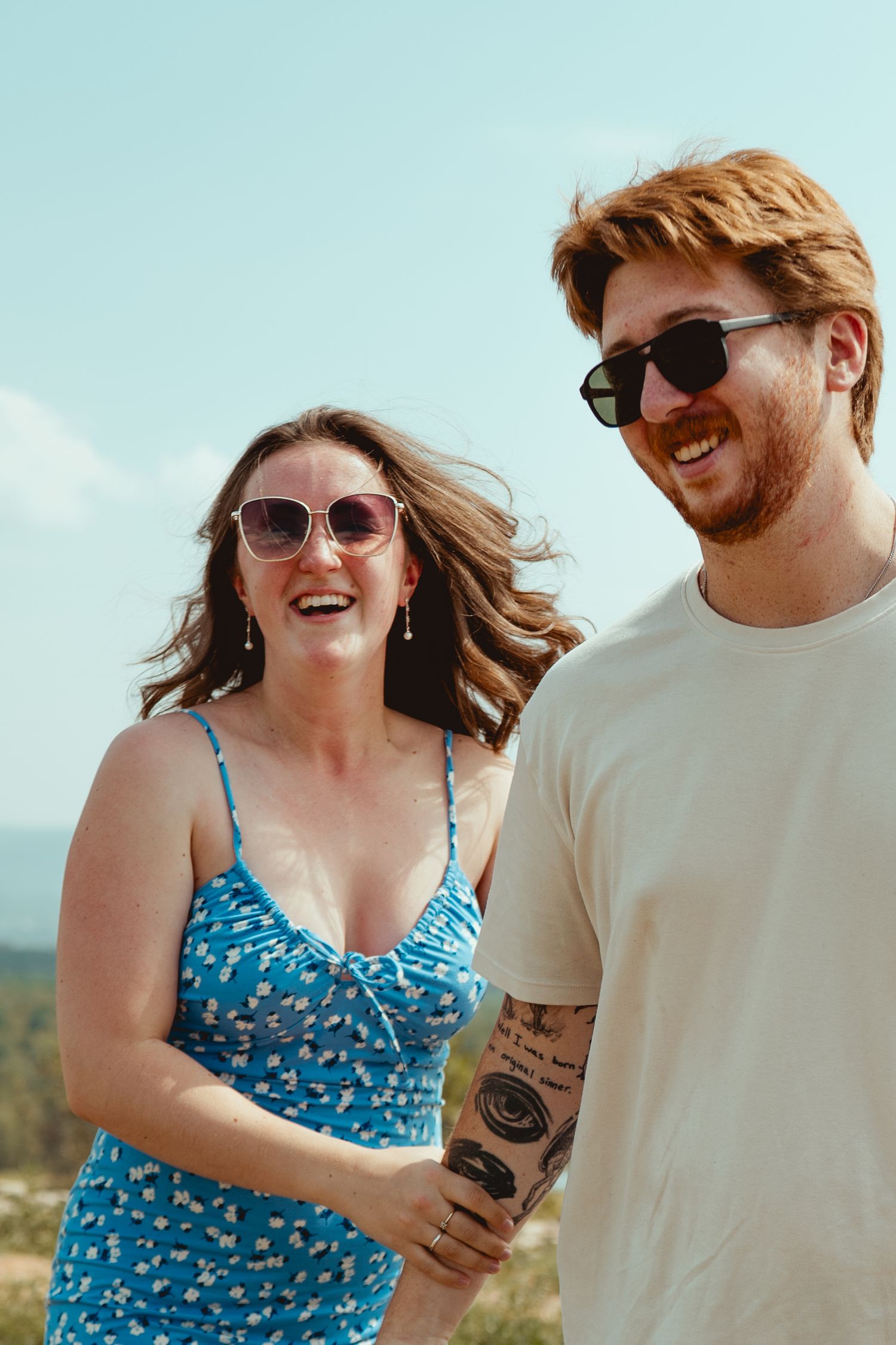 A couple shares joyful laughter while enjoying a sunny day outdoors, with one wearing a blue floral dress.