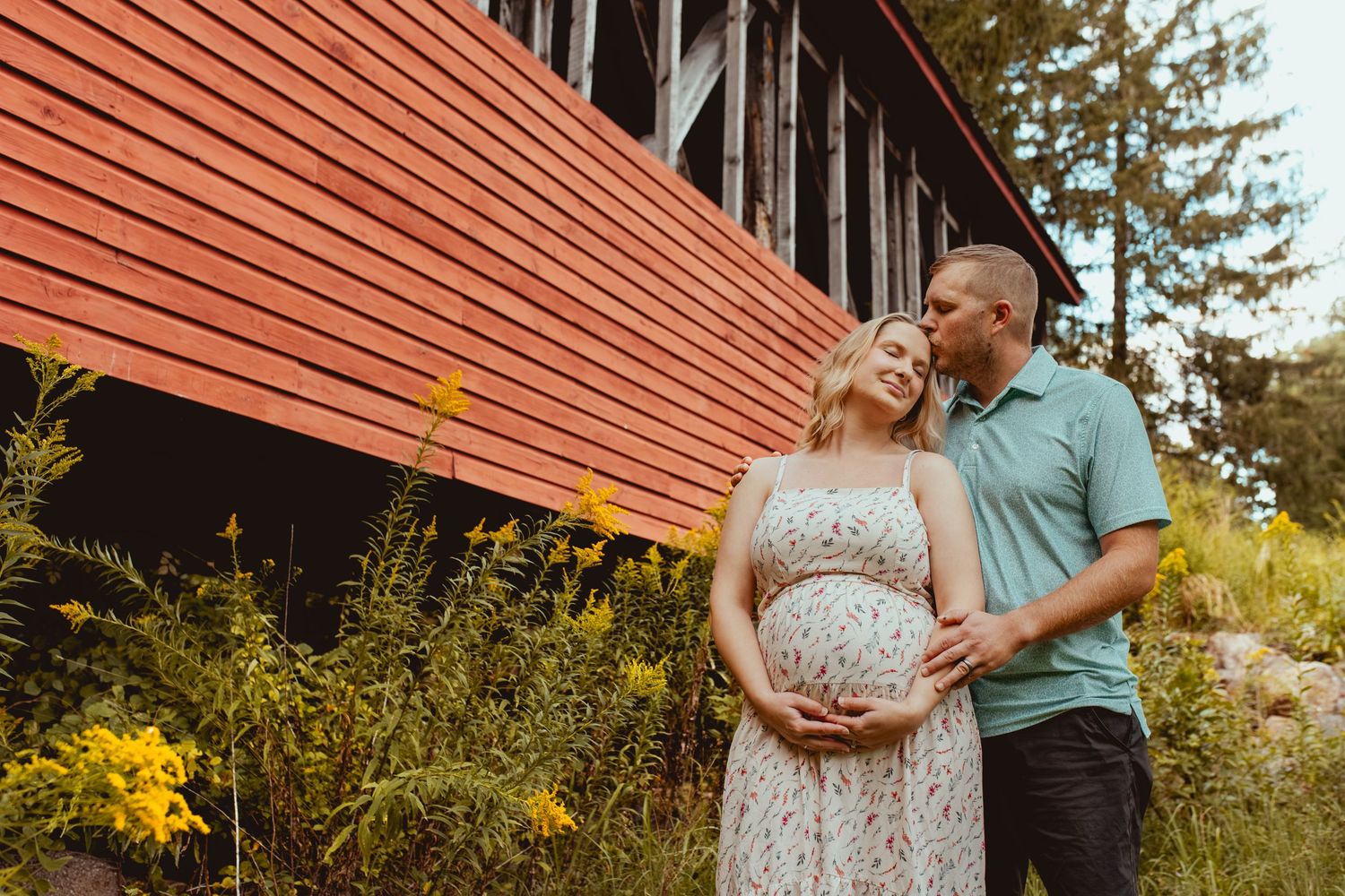 A couple embraces outdoors against rustic red siding and autumn foliage on a sunny day.