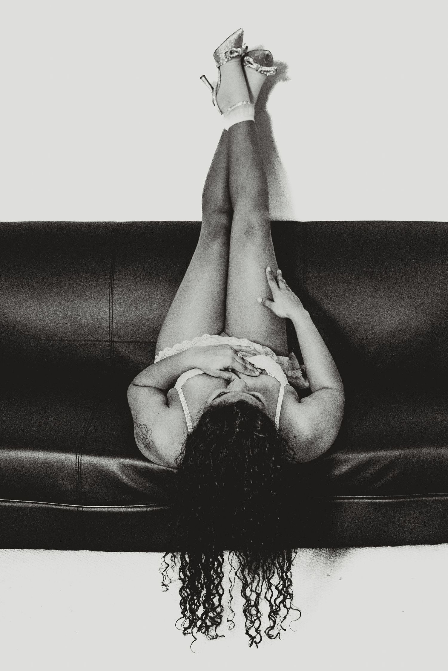 Black and white boudoir photo of a woman lying on a leather sofa with legs raised wearing heels.