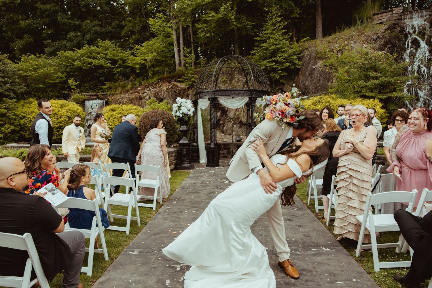 Groom dips bride for a romantic kiss at outdoor wedding ceremony with guests seated along the aisle.
