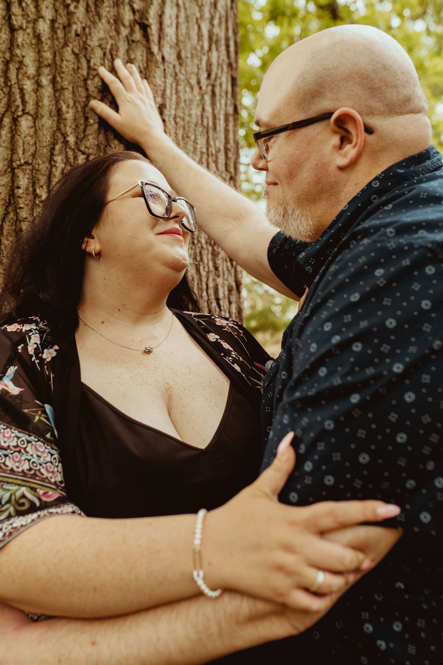 A couple shares a tender moment leaning against a tree, the man gently touching the womans hair while embracing her.