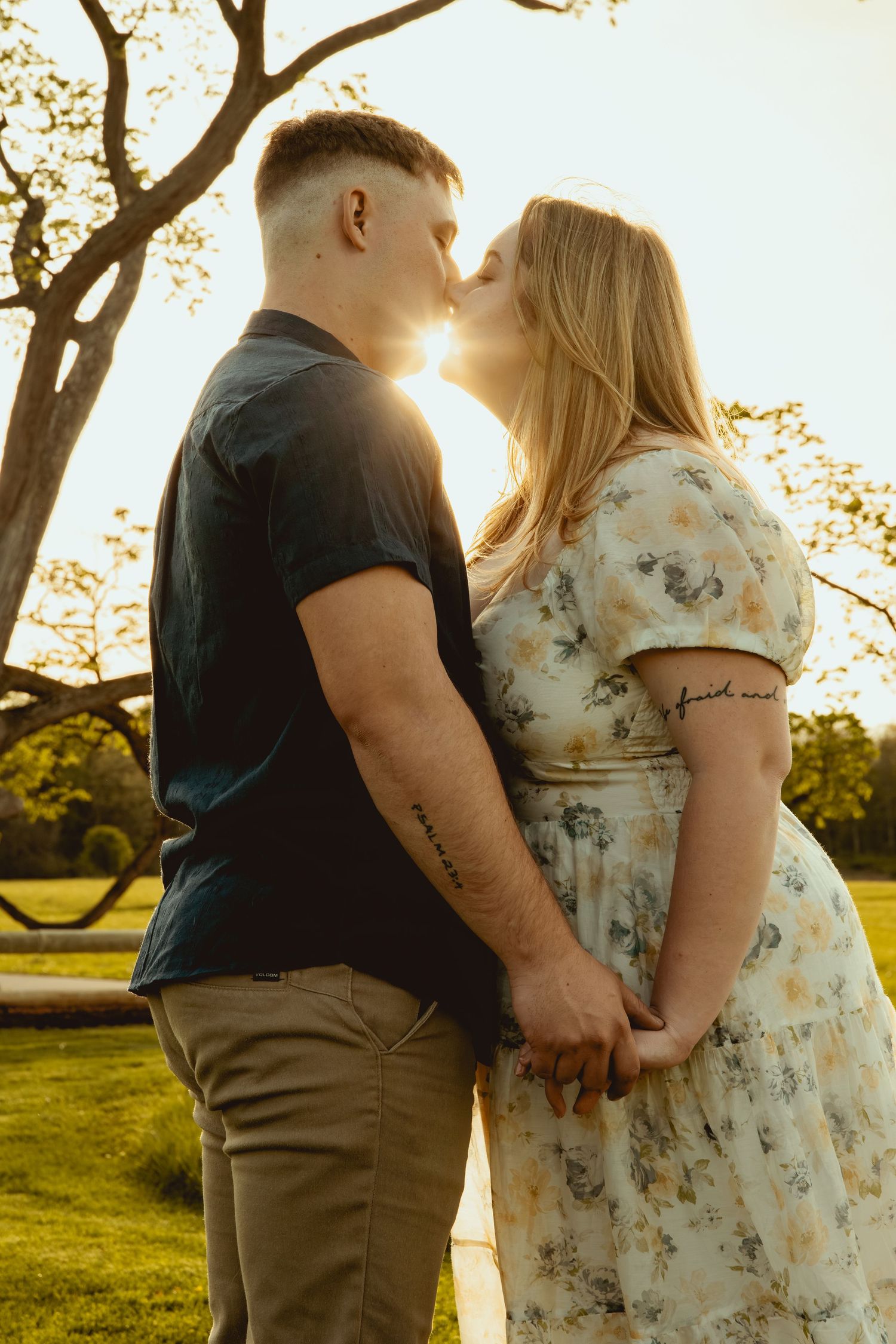 Couple sharing a kiss at golden hour in a park, holding hands, man in navy shirt, woman in floral dress.