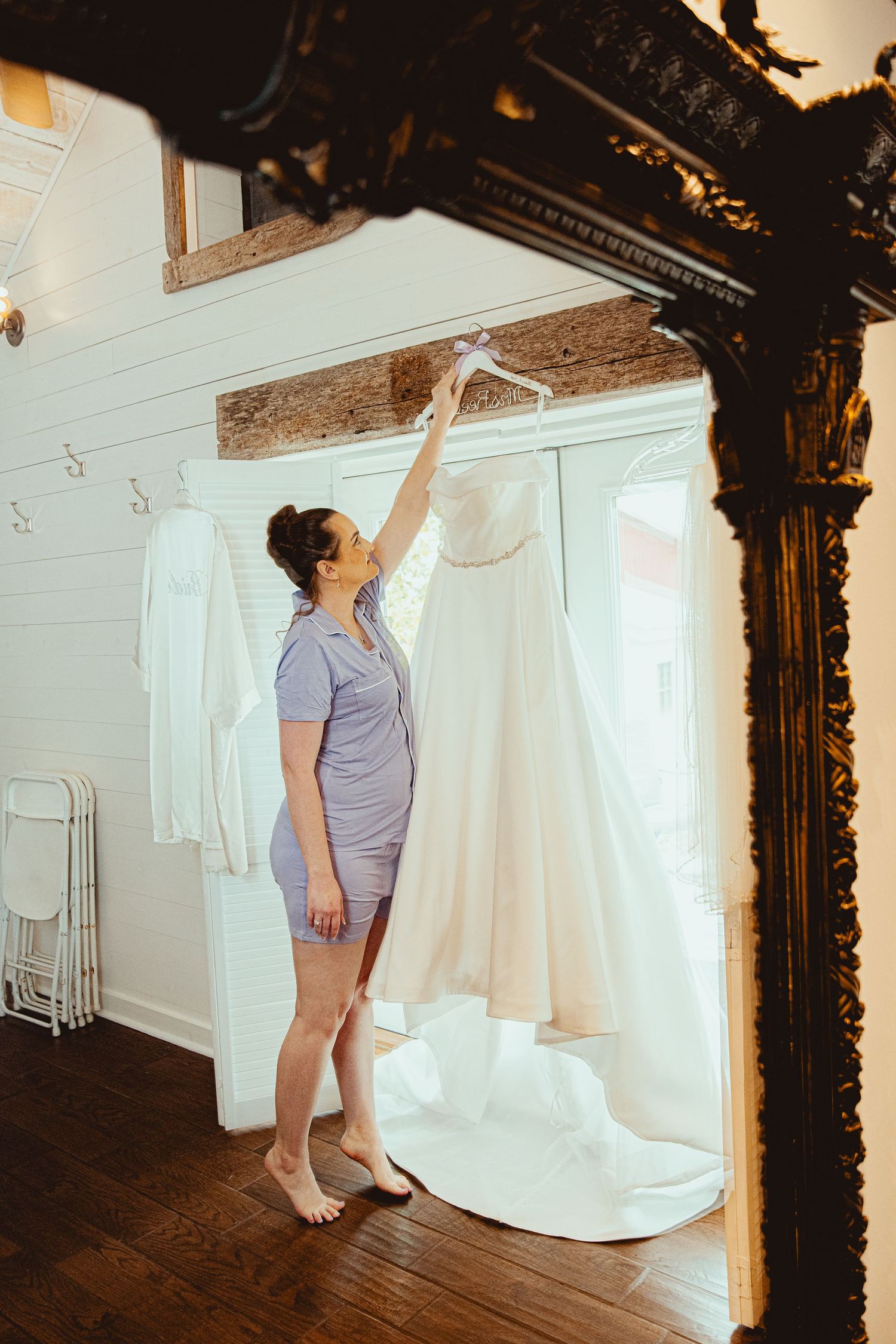 Bride reaching up to retrieve her wedding dress hanging in a rustic venue dressing room.
