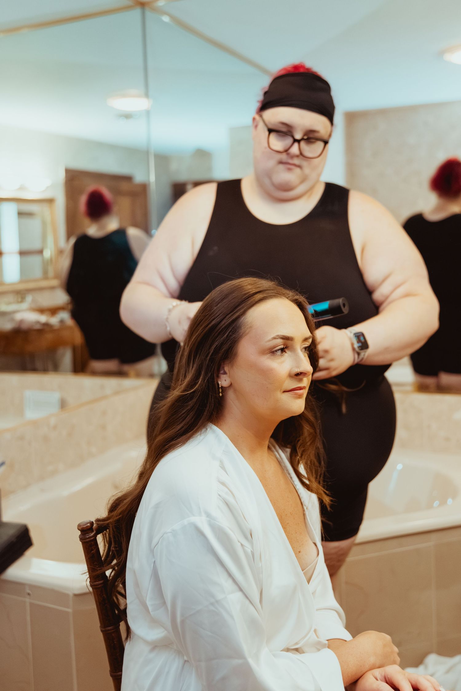 Hairstylist using curling iron on brunette bride in white robe, reflected in bathroom mirror.