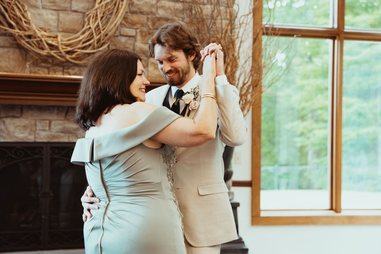 Groom in tan suit shares tender first dance with mother near stone fireplace and large windows at wedding reception.