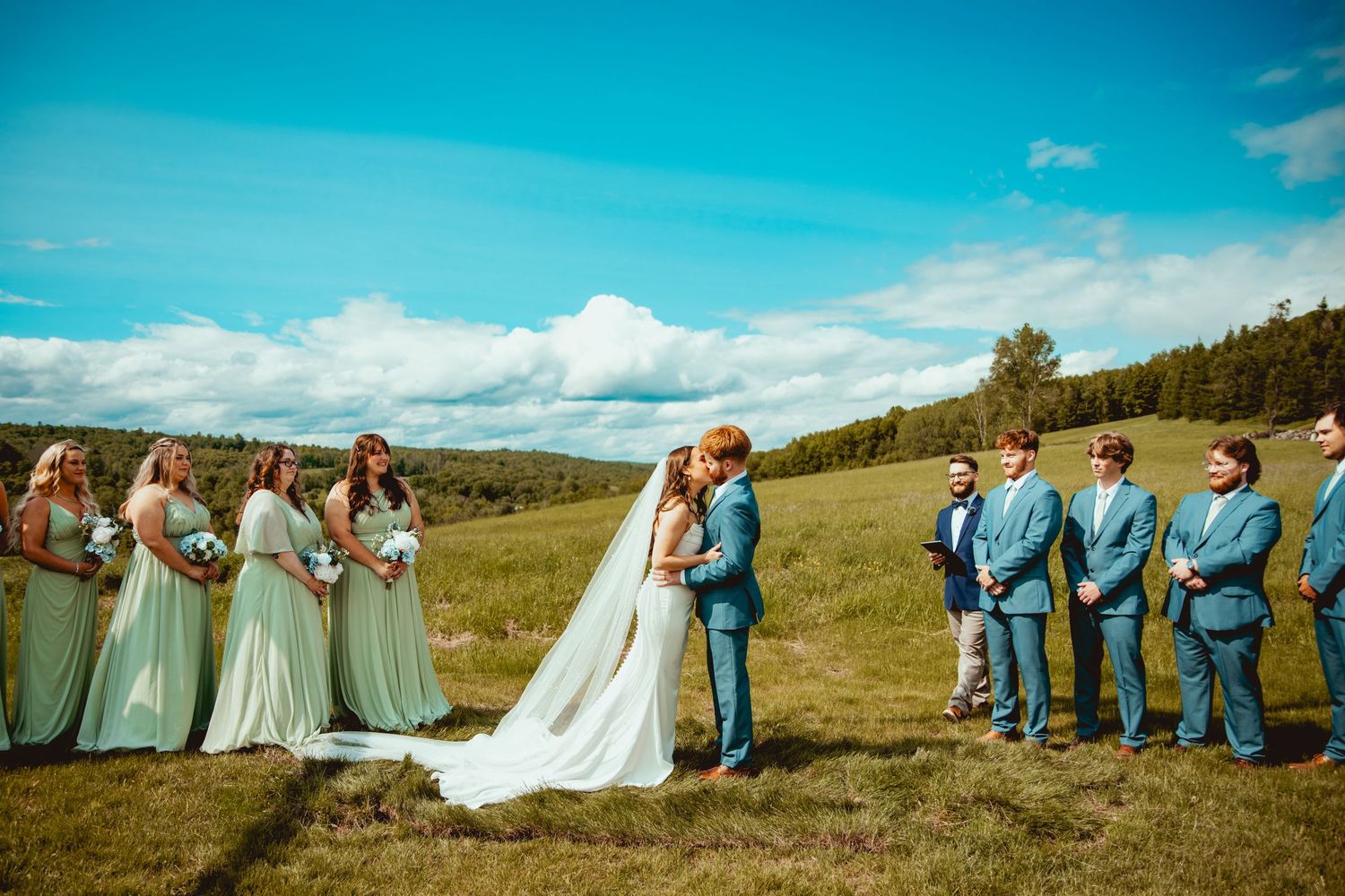 Wedding party creates fun photo composition on rolling green hillside under blue sky.