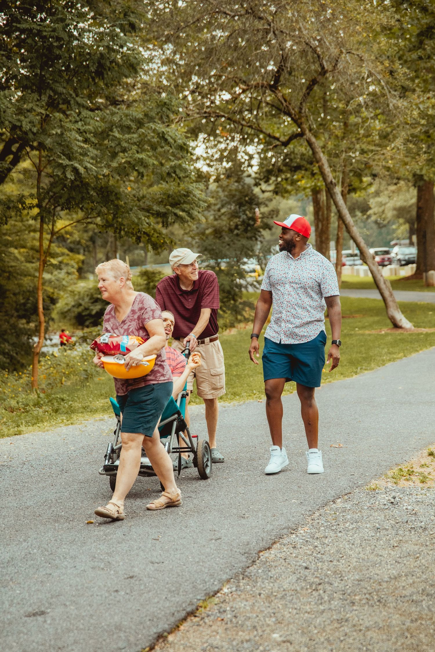 A group walks together on a gravel path through a wooded park area on a sunny afternoon.