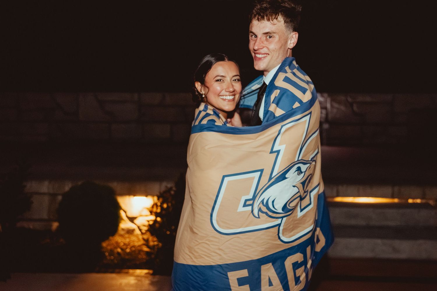 Person wrapped in a beige and blue blanket sits outside at night near glowing firelight.