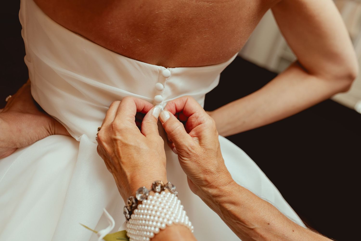 Hands fastening buttons on the back of a white wedding gown with pearl bracelet detail.