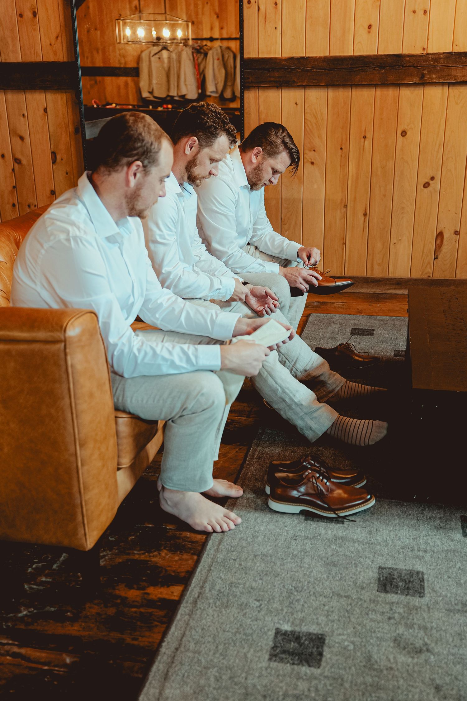 Groomsmen in white suits sitting together putting on brown dress shoes before wedding ceremony.