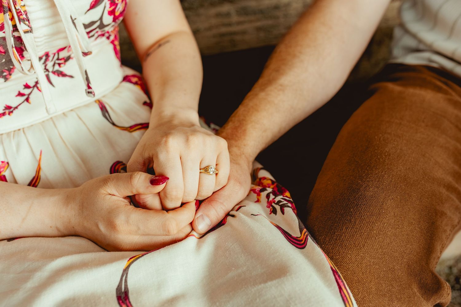 Close-up of a couple holding hands, woman wearing a gold ring and red nail polish, sitting together on a white dress.