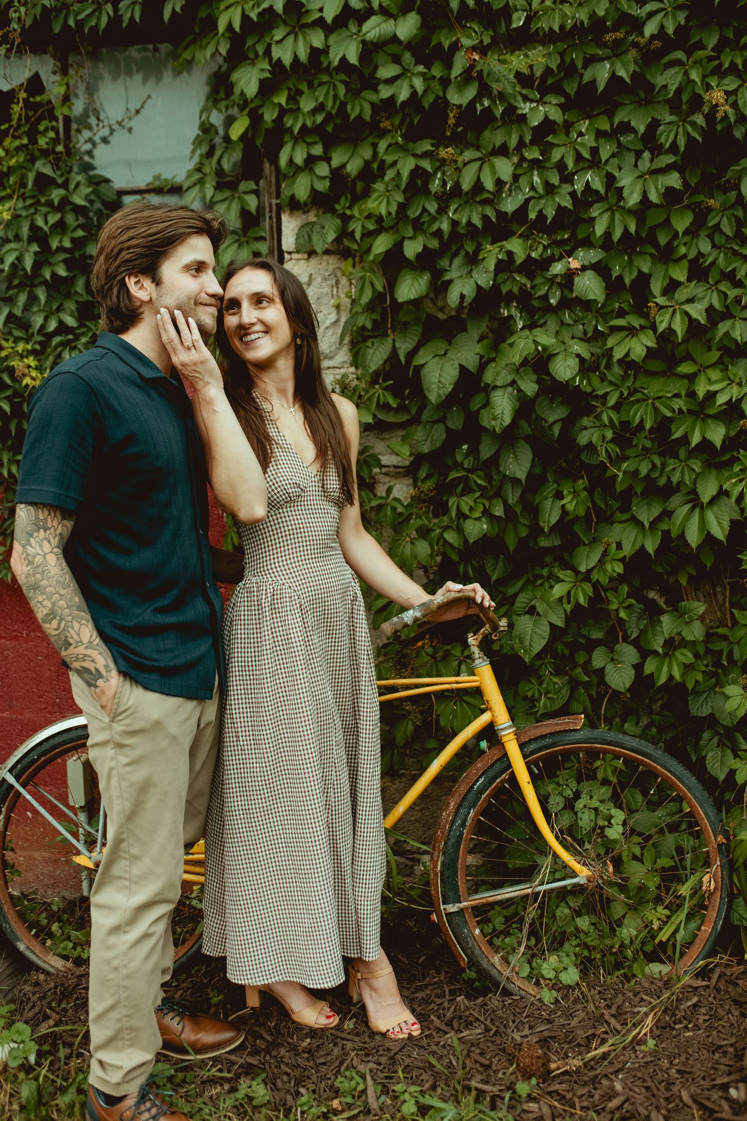 Couple posing by yellow bicycle against ivy-covered wall, man whispering to smiling woman in plaid dress.