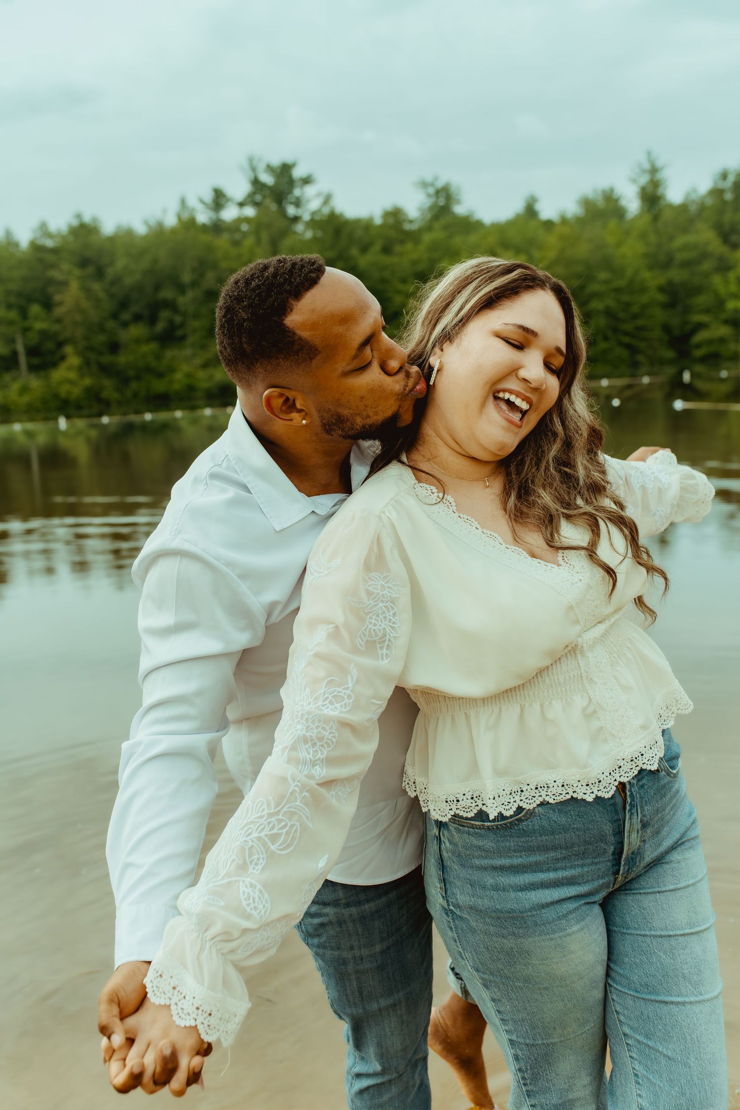 A joyful interracial couple shares a tender kiss by a lake, holding hands with lush green trees in the background.