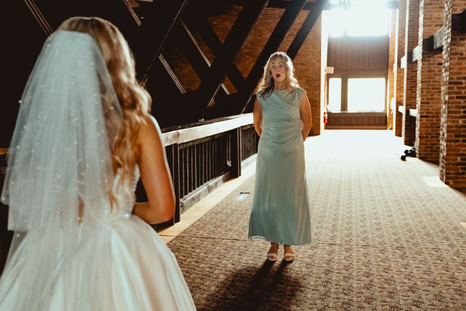 A bride in a white gown and veil shares an emotional first look moment with a woman in a teal dress indoors.
