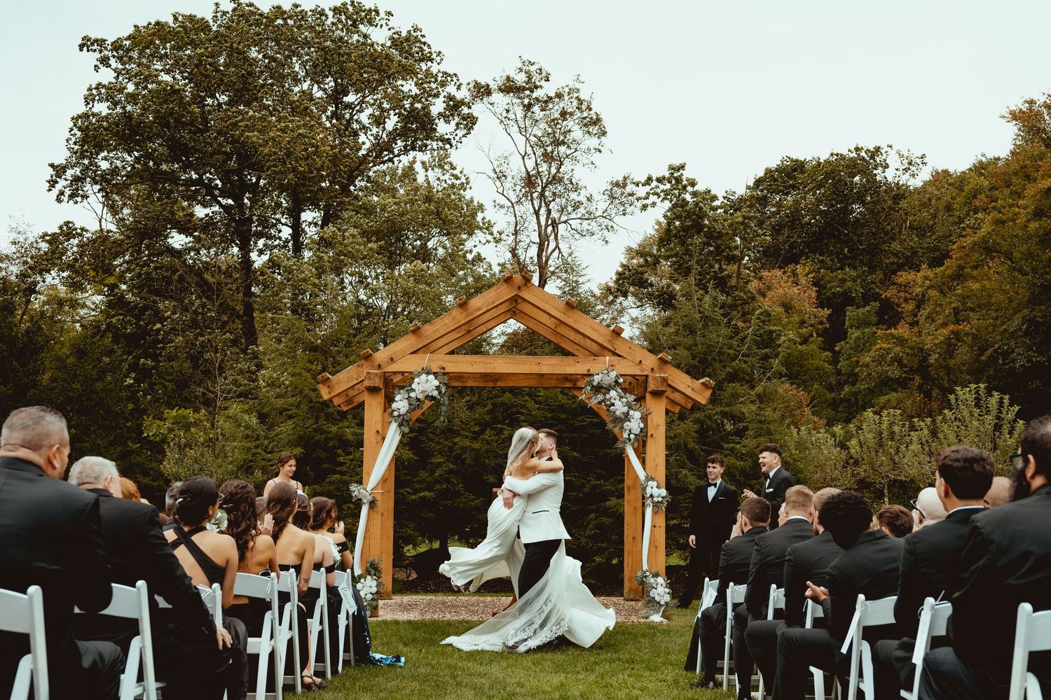 Bride and groom share first kiss under wooden arbor at outdoor wedding ceremony with white chairs.