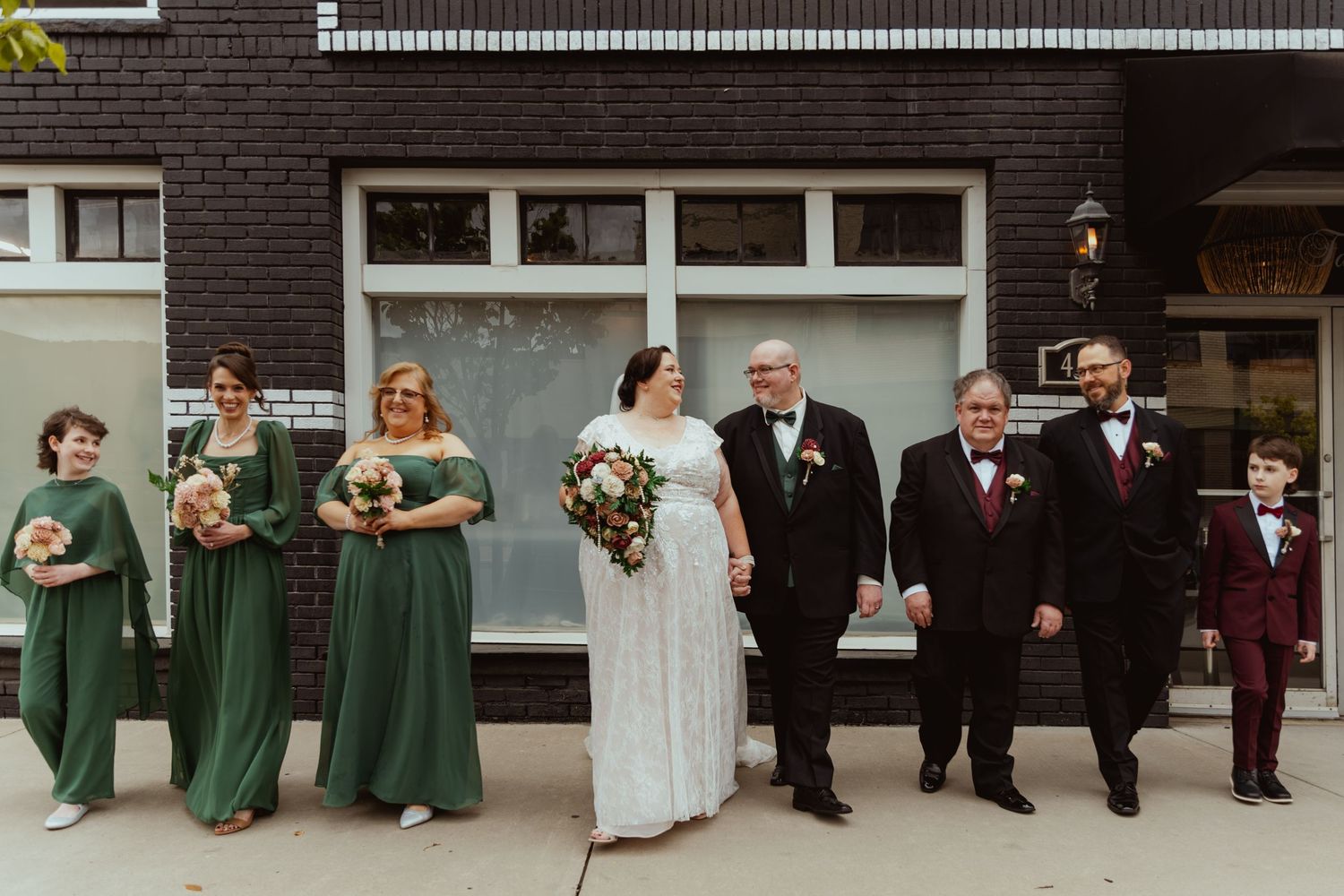 Bride in a white gown holding a bouquet stands with her groom, bridesmaids in green dresses, and groomsmen in dark suits outside a brick bui