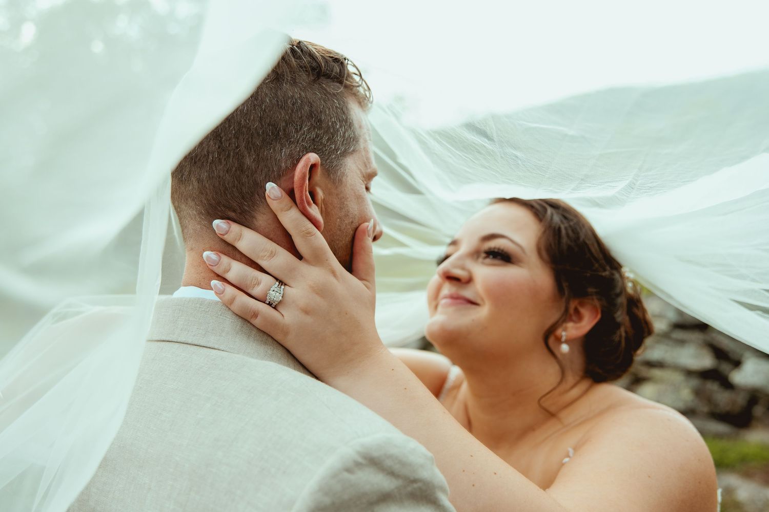 Bride and groom share intimate moment under flowing veil, bride smiling with wedding ring visible.