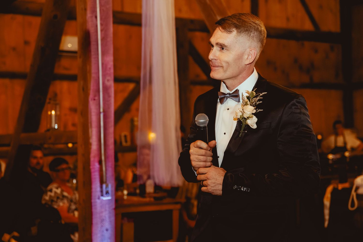 A speaker holds a microphone during a wedding reception in a warmly lit rustic barn venue.