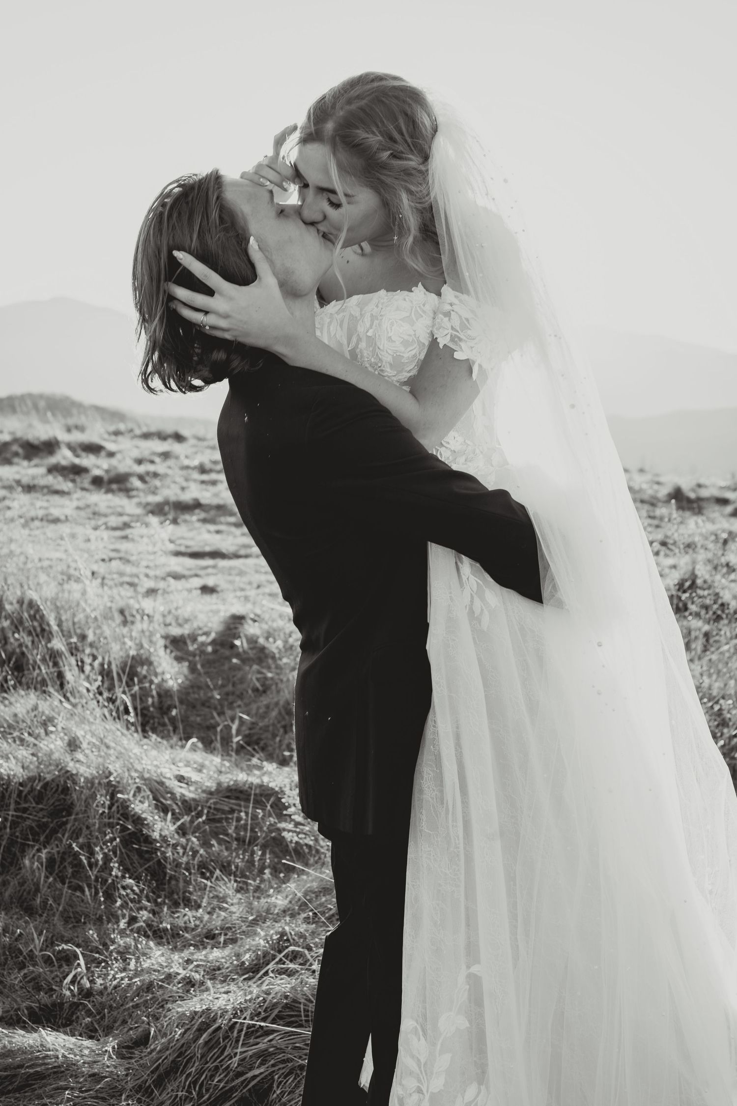 A romantic black and white wedding photo shows a couple embracing on a hilltop with the bride's veil flowing in the wind.
