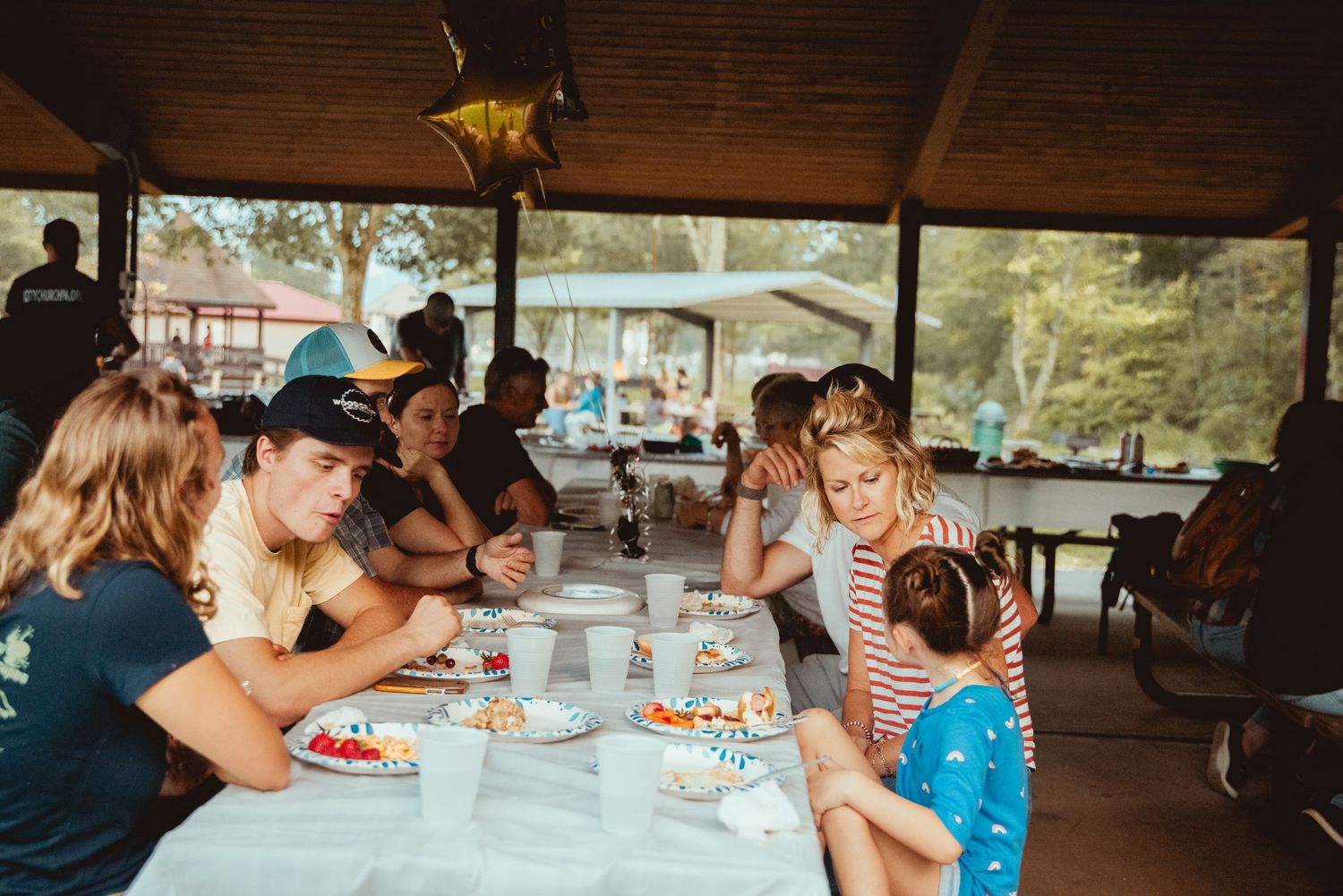 Group enjoying a meal together at a covered outdoor picnic pavilion on a sunny summer day.