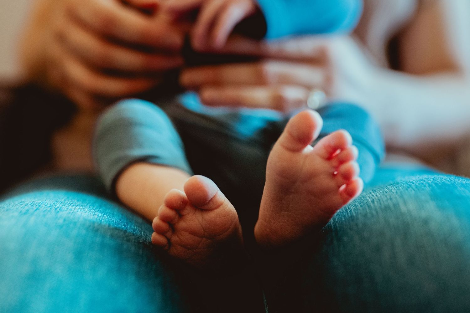 Small bare baby feet resting on turquoise fabric while hands hold a device in soft focus background.