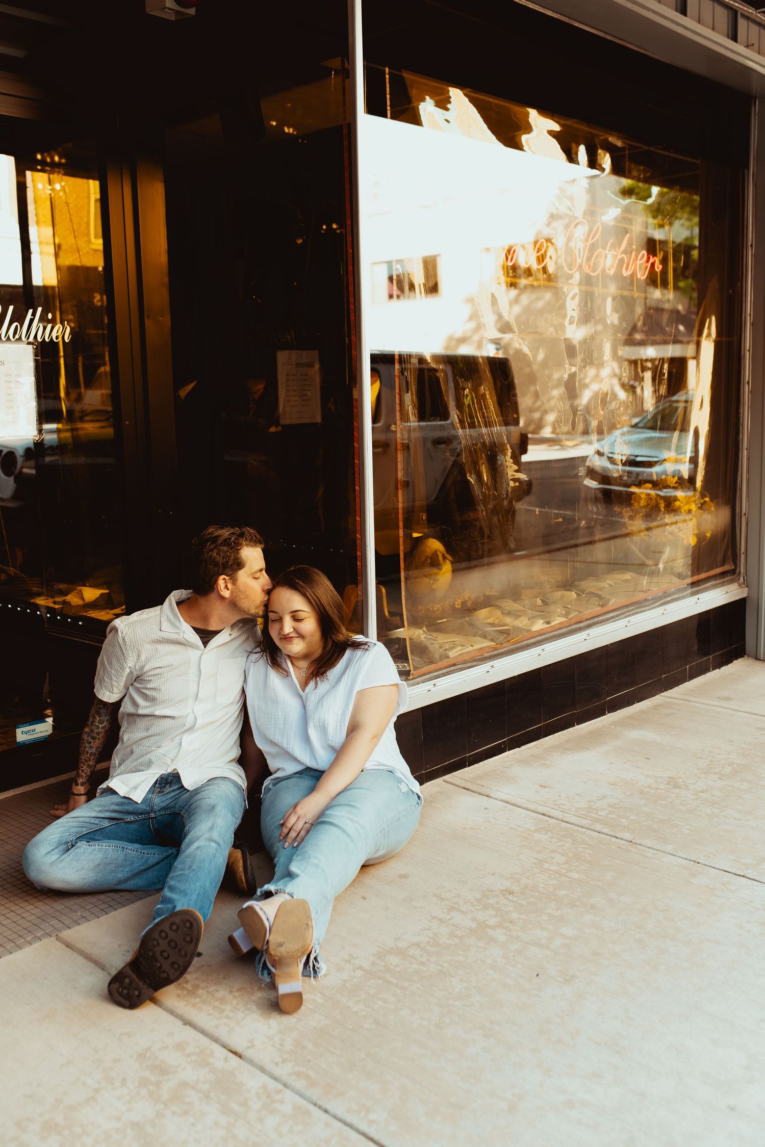 Couple sitting on sidewalk outside storefront, man kissing woman on cheek during golden hour engagement session.