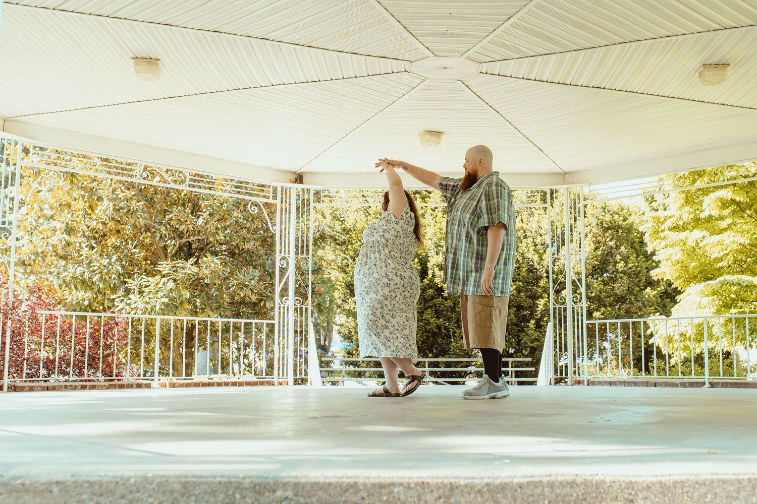 Couple dancing together under a white open-air pavilion surrounded by lush green trees on a sunny day.