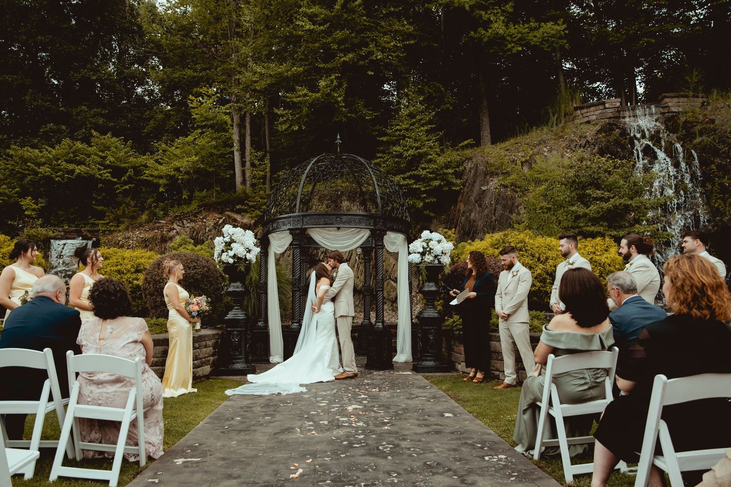 Outdoor wedding ceremony with couple at floral arch, guests seated on white chairs, waterfall backdrop in lush garden setting.