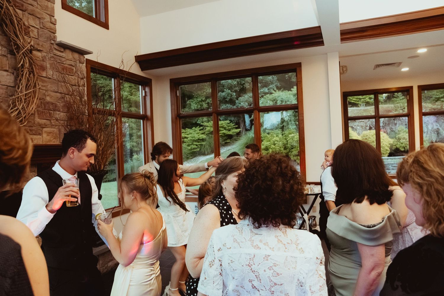 Wedding guests dance and mingle inside a rustic venue with large windows overlooking lush greenery.